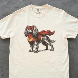 Peut inclure: T-shirt crème avec un dessin d'un chien déguisé en super-héros. Le chien porte un masque et une cape rouges, avec un emblème jaune. Le t-shirt est fait d'une matière douce et confortable.