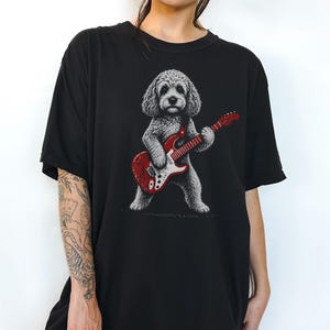 Könnte beinhalten: Ein schwarzes T-Shirt mit einer grafischen Illustration eines lockigen Hundes, der auf seinen Hinterbeinen steht und eine leuchtend rote E-Gitarre spielt. Der Hund ist in Grau- und Weißtönen auf dem einfarbigen schwarzen Stoff dargestellt.