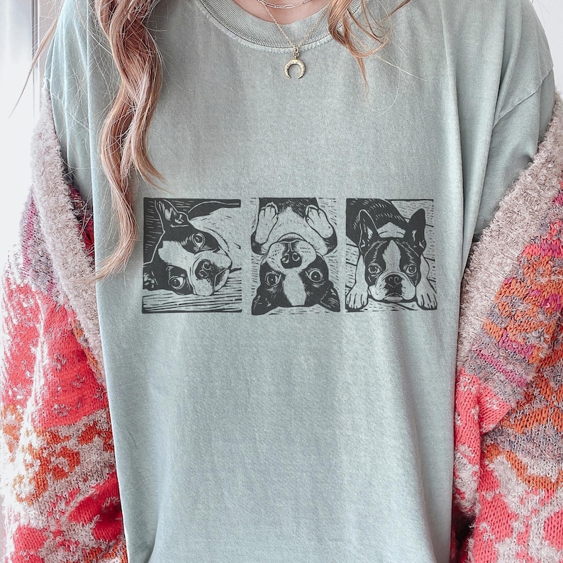 May include: A light green t-shirt featuring a black and white graphic of three Boston Terriers in different poses. The dogs are in rectangular frames, with one lying down, one upside down, and one looking forward.