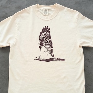 May include: Cream-colored t-shirt featuring a brown graphic of a hawk in flight. The bird's wings are spread wide, showcasing detailed feather patterns. The shirt has a classic crew neck and short sleeves, suitable for casual wear.
