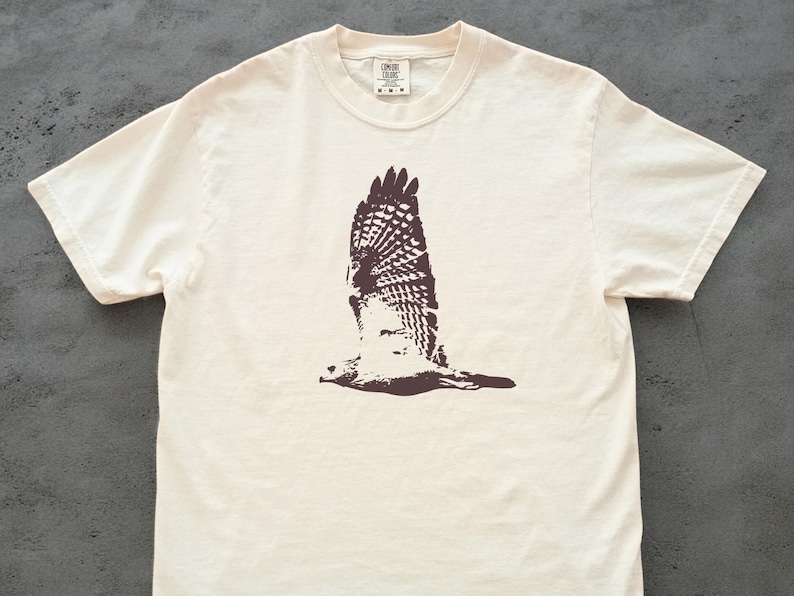 May include: Cream-colored t-shirt featuring a brown graphic of a hawk in flight. The bird's wings are spread wide, showcasing detailed feather patterns. The shirt has a classic crew neck and short sleeves, suitable for casual wear.