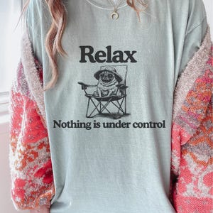 May include: A pale blue t-shirt featuring the word "Relax" above a graphic of a pug in a hat, seated in a folding chair. The text below the graphic reads "Nothing is under control."