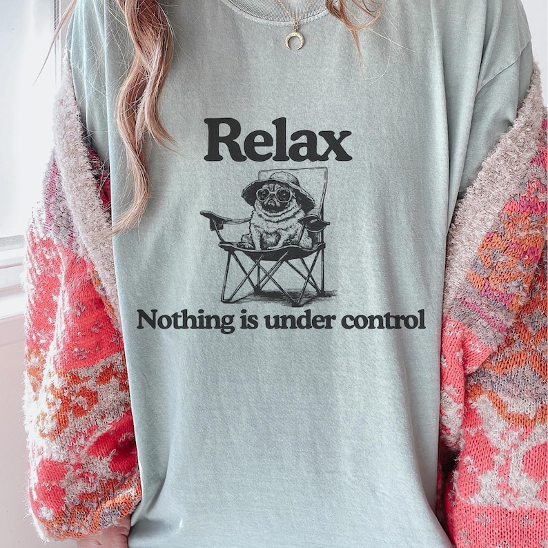 May include: A pale blue t-shirt featuring the word "Relax" above a graphic of a pug in a hat, seated in a folding chair. The text below the graphic reads "Nothing is under control."