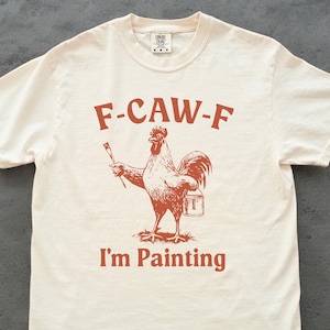 May include: Cream-colored t-shirt with a rust-colored rooster graphic holding a paintbrush and paint bucket. The text "F-CAW-F" is above the rooster, and "I'm Painting" is below. The shirt has a crew neck and short sleeves.
