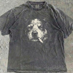 May include: A dark grey t-shirt with a graphic of a dog smoking a cigarette. The dog's face is white against the grey fabric. The t-shirt has a crew neck and short sleeves. The fabric has a distressed appearance.
