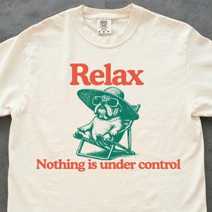 May include: Cream-colored t-shirt with a graphic of a bulldog wearing sunglasses and a hat, relaxing in a beach chair. The word "Relax" is above the dog, and "Nothing is under control" is below.