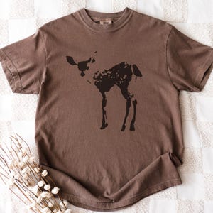 May include: Brown t-shirt featuring a dark brown silhouette of a deer. The deer has a spotted pattern on its body. The shirt is made of a soft material and has short sleeves. The shirt is laying on a white and cream checkered surface.