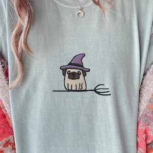 May include: Pale blue t-shirt with an embroidered pug wearing a purple witch's hat and sitting on a broom. The design is in the centre of the shirt. The shirt is made of a soft, comfortable material.