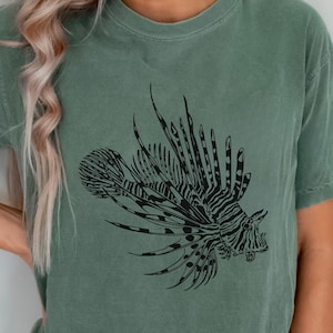 Op de afbeelding: Saliegroen t-shirt met een gedetailleerde zwarte illustratie van een koraalduivel. Het koraalduivelontwerp is gecentreerd en toont zijn kenmerkende vinnen en strepen. Het shirt heeft een klassieke ronde hals en korte mouwen.