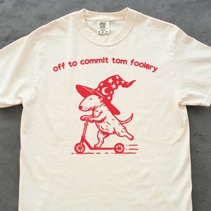 May include: Cream-colored t-shirt with a red graphic of a dog riding a scooter and wearing a wizard hat. The text "off to commit tom foolery" is arched above the graphic in red. The shirt has a crew neck and short sleeves.