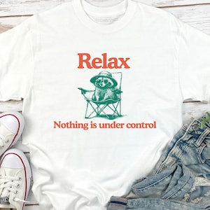 May include: White t-shirt with the word "Relax" in orange above a green illustration of a raccoon wearing sunglasses and a hat, sitting in a folding chair. Below the image is the phrase "Nothing is under control".