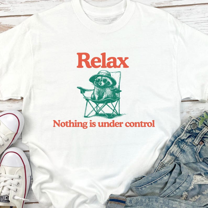 May include: White t-shirt with the word "Relax" in orange above a green illustration of a raccoon wearing sunglasses and a hat, sitting in a folding chair. Below the image is the phrase "Nothing is under control".