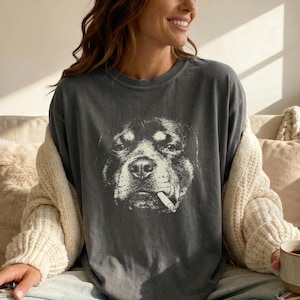 Könnte beinhalten: Dunkelgraues T-Shirt mit einer schwarz-weißen Grafik eines Hundes mit Zigarette im Maul. Das Gesicht des Hundes ist der Mittelpunkt, mit detaillierten Schattierungen. Das T-Shirt hat eine lockere Passform.