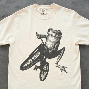Pu&ograve; includere: T-shirt color crema con un'illustrazione in bianco e nero di una rana in bicicletta. La rana &egrave; dettagliata e la bicicletta ha sottili raggi. La maglietta ha un girocollo e maniche corte. La maglietta &egrave; in materiale morbido.