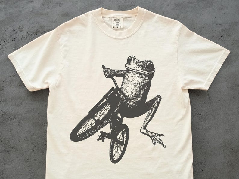 Pu&ograve; includere: T-shirt color crema con un'illustrazione in bianco e nero di una rana in bicicletta. La rana &egrave; dettagliata e la bicicletta ha sottili raggi. La maglietta ha un girocollo e maniche corte. La maglietta &egrave; in materiale morbido.