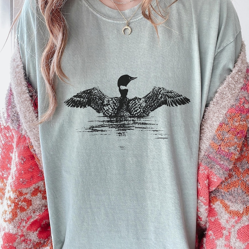 May include: A light green t-shirt featuring a black and white illustration of a loon with outstretched wings on water. The loon is centered on the shirt, with the water depicted as horizontal lines. The shirt has a crew neck.