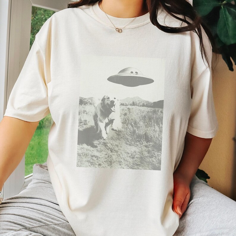 May include: A white t-shirt with a graphic print of a dog looking up at a flying saucer. The dog is in a field of grass.