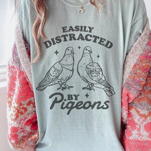 May include: A pale blue t-shirt featuring the text "EASILY DISTRACTED BY PIGEONS" and two pigeon illustrations. The shirt has a crew neck and short sleeves. The graphic is in black.