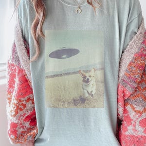 May include: A light blue t-shirt with a graphic of a corgi running towards a flying saucer. The t-shirt has a fuzzy, orange and white cardigan over it.