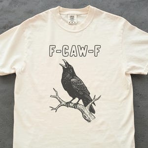 May include: Cream-colored t-shirt featuring a black illustration of a crow perched on a branch, with the text "F-CAW-F" above. The crow is depicted with its beak open, as if cawing. The shirt has a classic crew neck and short sleeves.
