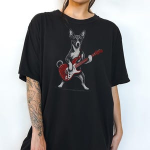 May include: A black short-sleeve t-shirt featuring a graphic illustration of a dog standing on its hind legs while playing a bright red electric guitar. The dog is rendered in shades of black, white, and gray, contrasting against the solid black fabric.