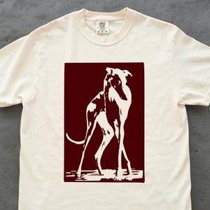 以下が含まれることがあります： クリーム色のTシャツ。濃い赤色の長方形の中に、白い犬のグラフィックが描かれています。犬は細長い脚と曲がった尾を持つ、ミニマルなデザインです。Tシャツはクルーネックです。