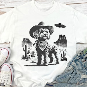 Pu&ograve; includere: T-shirt bianca con grafica in bianco e nero di un cane con cappello da cowboy e bandana, ambientato in un paesaggio desertico con cactus e montagne. Un UFO &egrave; raffigurato sopra il cane.