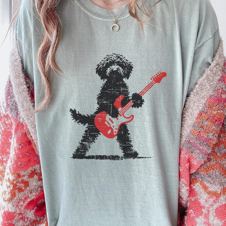 May include: A light green t-shirt featuring a black dog playing a red electric guitar. The dog is silhouetted with a distressed texture. The guitar has white accents. The shirt is a comfortable, casual style.