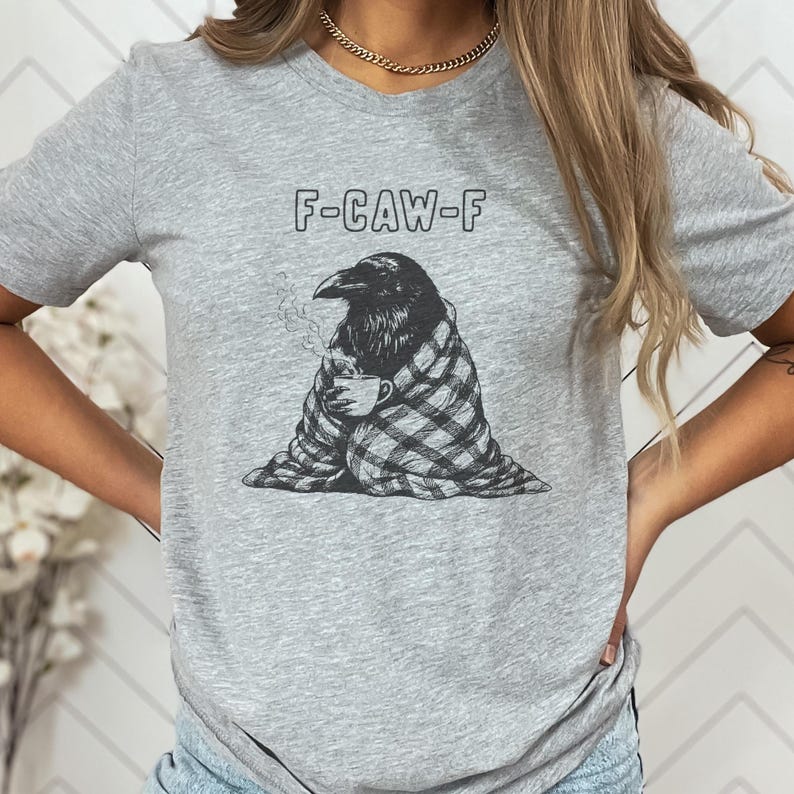 May include: A gray t-shirt featuring a black and white graphic of a raven wrapped in a plaid blanket, holding a mug with steam rising. The text "F-CAW-F" is above the raven. The shirt is a casual style.