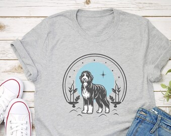 Może przedstawiać: Szary melanżowy t-shirt z krótkim rękawem z nadrukiem graficznym przedstawiającym czarno-białego psa na tle jasnoniebieskiego koła. Wzór zawiera ciemnoszarą łukowatą ramkę, małe gwiazdki oraz stylizowane elementy roślinne po obu stronach.