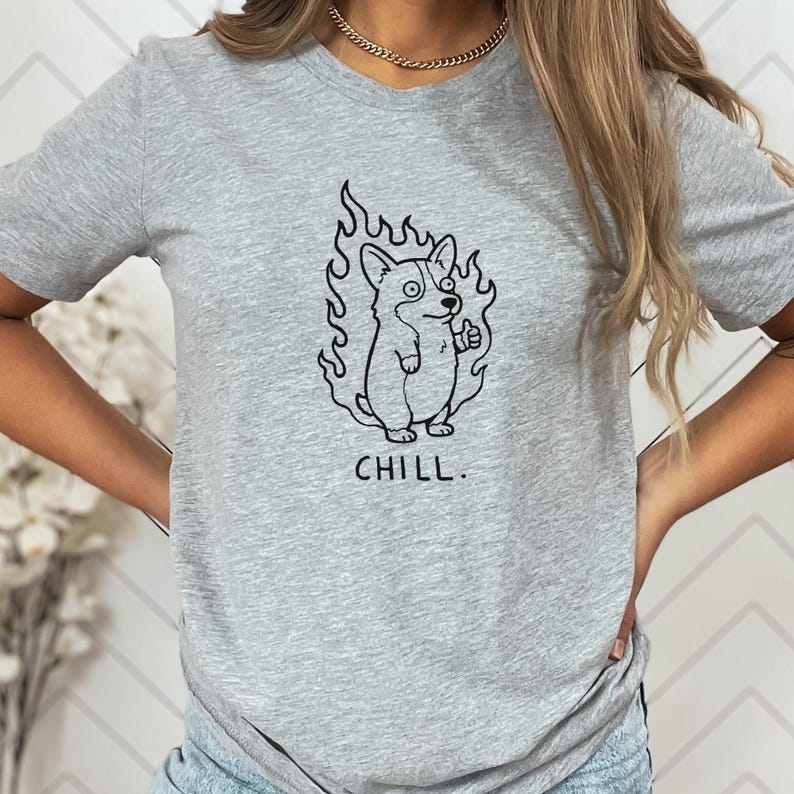 May include: A heather gray t-shirt featuring a black graphic of a corgi dog surrounded by flames, giving a thumbs-up. The word "CHILL" is printed below the graphic. The shirt is a casual wear item.