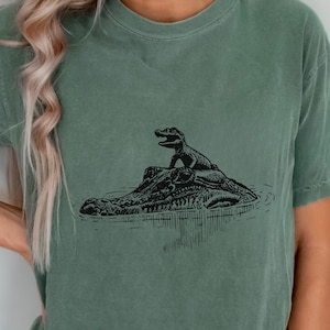 Op de afbeelding: Saliegroen t-shirt met een zwarte illustratie van een alligator en een kleinere alligator op zijn rug, in water. Het ontwerp is gecentreerd op het shirt.