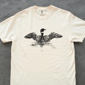 May include: Cream-colored t-shirt featuring a black and white illustration of a loon with outstretched wings, appearing to float on water. The shirt has a classic crew neck and short sleeves, with a small tag at the neckline.