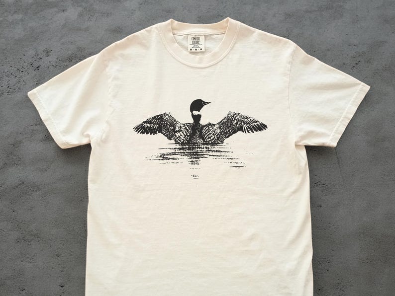 May include: Cream-colored t-shirt featuring a black and white illustration of a loon with outstretched wings, appearing to float on water. The shirt has a classic crew neck and short sleeves, with a small tag at the neckline.