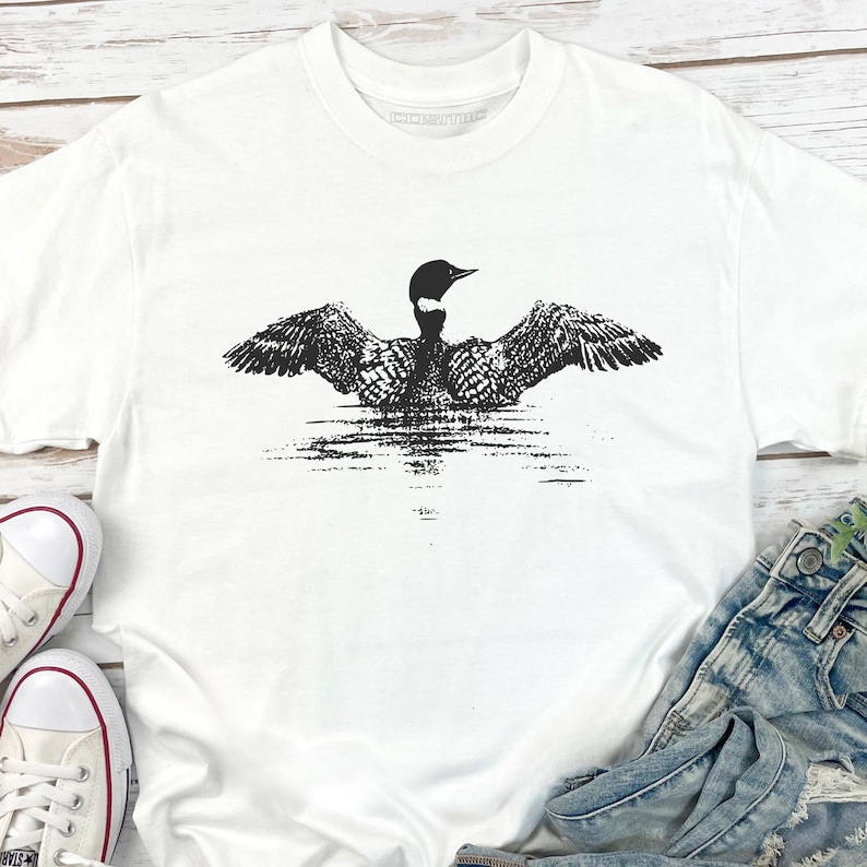 May include: White t-shirt featuring a black and white graphic of a loon with outstretched wings on a body of water. The bird's wings are detailed with intricate patterns. The shirt is made of a soft material.