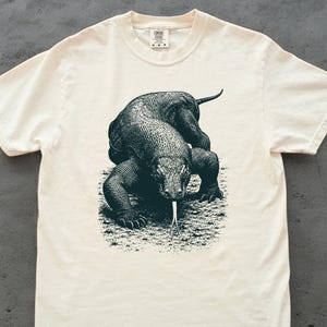 Può includere: Una t-shirt a maniche corte color crema con una dettagliata stampa grafica nera di un drago di Komodo sul davanti. La lucertola è raffigurata in una posizione bassa e furtiva con la lingua biforcuta estesa. Girocollo ed etichetta interna.