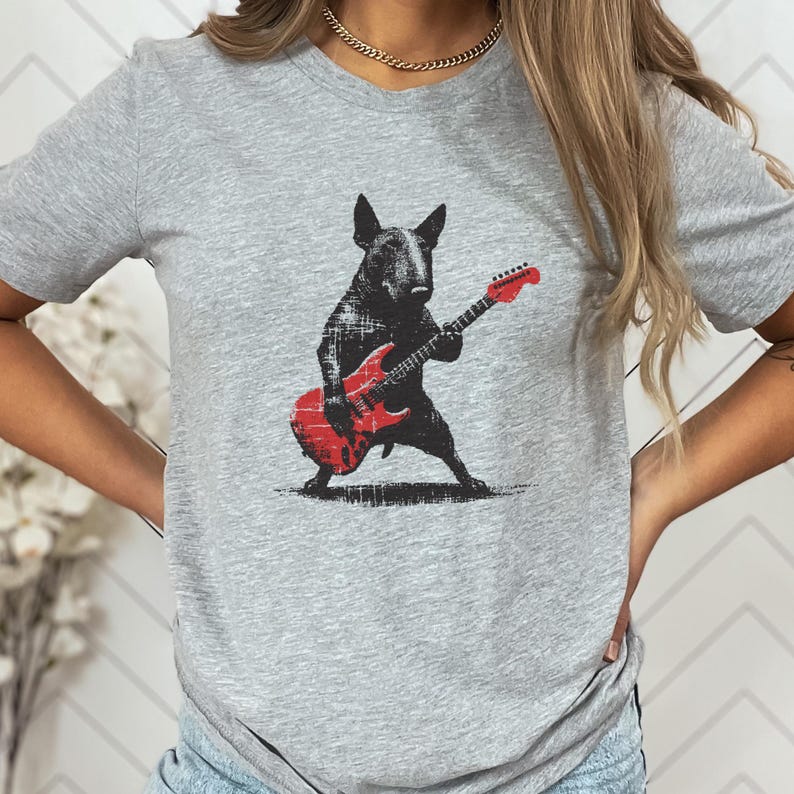 K&ouml;nnte beinhalten: Ein graues T-Shirt mit einem schwarzen Hund, der eine rote E-Gitarre spielt. Der Hund ist eine stilisierte Illustration mit einem Used-Look im Vintage-Stil. Die Gitarre hat eine klassische Form. Das Shirt ist ein l&auml;ssiges Kleidungsst&uuml;ck f&uuml;r den Alltag.