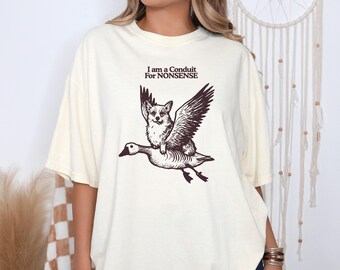 Könnte beinhalten: Cremefarbenes T-Shirt mit einer skurrilen Illustration eines Corgis, der auf einer Gans mit ausgebreiteten Flügeln reitet. Der Text "I am a Conduit For NONSENSE" ist über dem Bild gedruckt. Das Shirt hat eine lockere Passform.