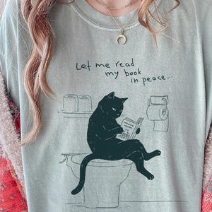 Puede incluir: Camiseta verde claro con un gato negro leyendo un libro en un inodoro. El texto "Let me read my book in peace..." está encima del gato. La camiseta tiene un corte holgado.