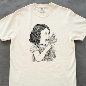 May include: Cream-colored t-shirt featuring a black and white illustration of a young girl holding a praying mantis. The girl is depicted with her mouth open in surprise. The t-shirt is made of a soft, comfortable material.