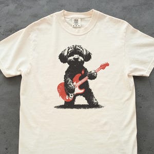 Può includere: T-shirt color crema con un cane nero che suona una chitarra elettrica rossa. Il cane ha un aspetto ispido e la chitarra ha dettagli bianchi. Il design è stampato su una maglietta morbida e casual.