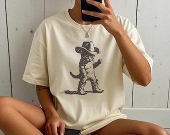 May include: Cream-colored t-shirt featuring a sepia-toned illustration of a cat dressed as a cowboy. The cat wears a cowboy hat and boots, with one arm raised. The shirt has a relaxed fit and short sleeves.