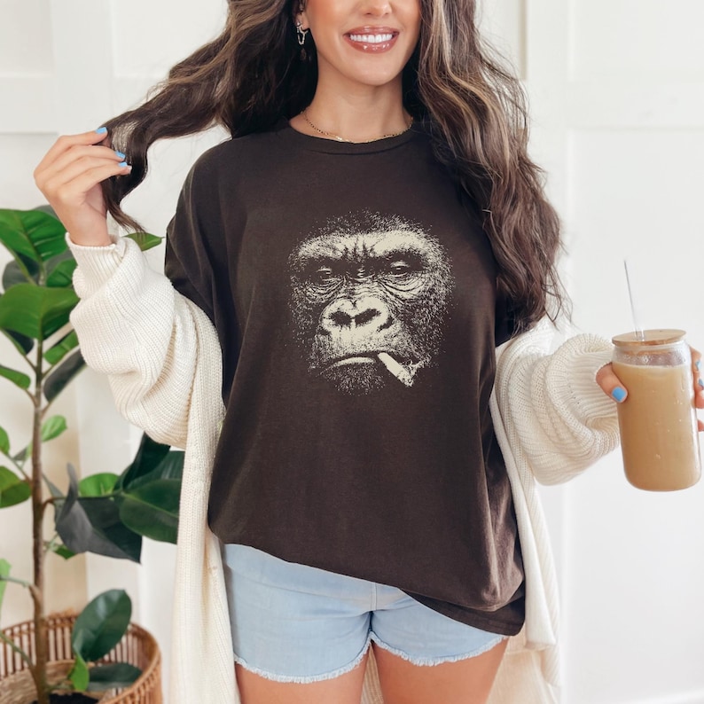 May include: A dark brown t-shirt featuring a detailed graphic of a gorilla smoking a cigarette. The gorilla's face is rendered in a vintage, distressed style. The shirt is a relaxed fit, suitable for casual wear.