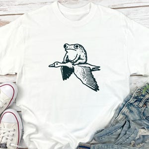 K&ouml;nnte beinhalten: Ein wei&szlig;es kurz&auml;rmeliges T-Shirt mit einem schwarzen Grafikdruck, der einen Frosch auf dem R&uuml;cken einer fliegenden Gans zeigt. Die Illustration ist mittig auf der Brust des Kleidungsst&uuml;cks platziert und zeigt einen detaillierten Strichzeichnungsstil.