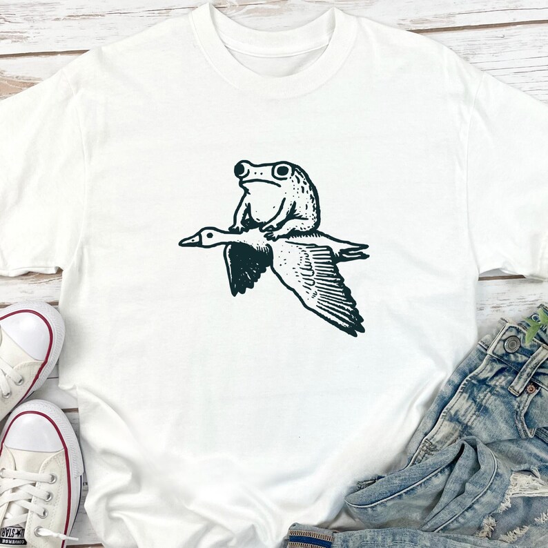 K&ouml;nnte beinhalten: Ein wei&szlig;es kurz&auml;rmeliges T-Shirt mit einem schwarzen Grafikdruck, der einen Frosch auf dem R&uuml;cken einer fliegenden Gans zeigt. Die Illustration ist mittig auf der Brust des Kleidungsst&uuml;cks platziert und zeigt einen detaillierten Strichzeichnungsstil.