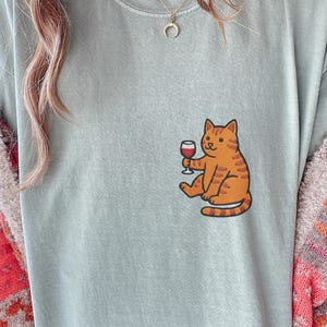 May include: A light blue t-shirt featuring an orange tabby cat illustration holding a glass of red wine. The cat is sitting, and the design is on the front of the shirt. The shirt has a relaxed fit.