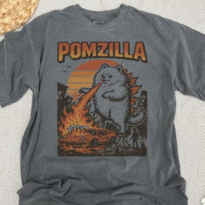 May include: Dark grey t-shirt featuring the word "POMZILLA" in orange and brown. The graphic shows a Pomeranian dog as Godzilla, breathing fire onto a car and buildings. A person and dog are in the background.