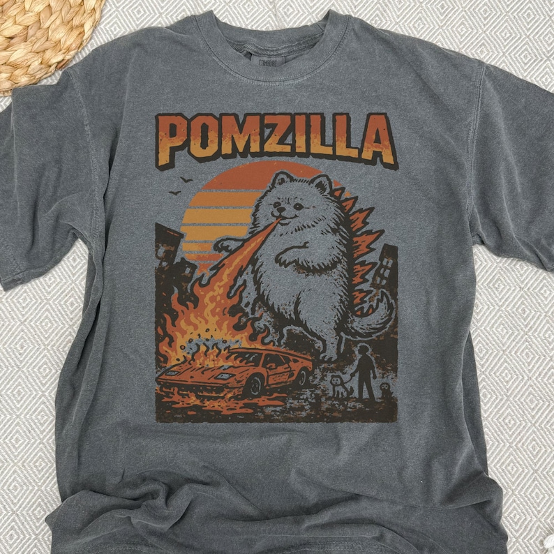 May include: Dark grey t-shirt featuring the word "POMZILLA" in orange and brown. The graphic shows a Pomeranian dog as Godzilla, breathing fire onto a car and buildings. A person and dog are in the background.