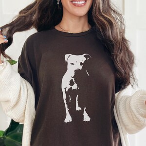 Pode incluir: Camiseta marrom escuro com um gr&aacute;fico branco de um cachorro. O cachorro est&aacute; sentado. A camiseta &eacute; de cor s&oacute;lida e tem gola redonda. A modelo est&aacute; usando um cardigan branco.