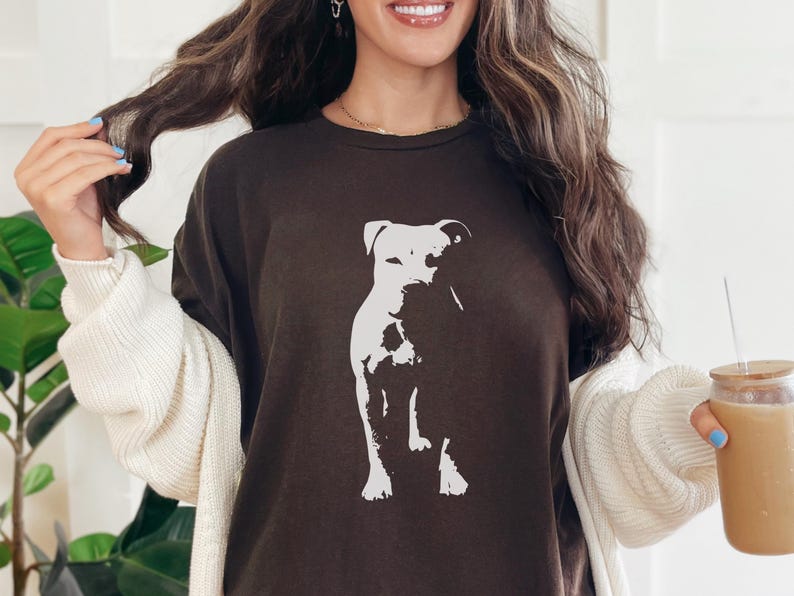 Pode incluir: Camiseta marrom escuro com um gr&aacute;fico branco de um cachorro. O cachorro est&aacute; sentado. A camiseta &eacute; de cor s&oacute;lida e tem gola redonda. A modelo est&aacute; usando um cardigan branco.